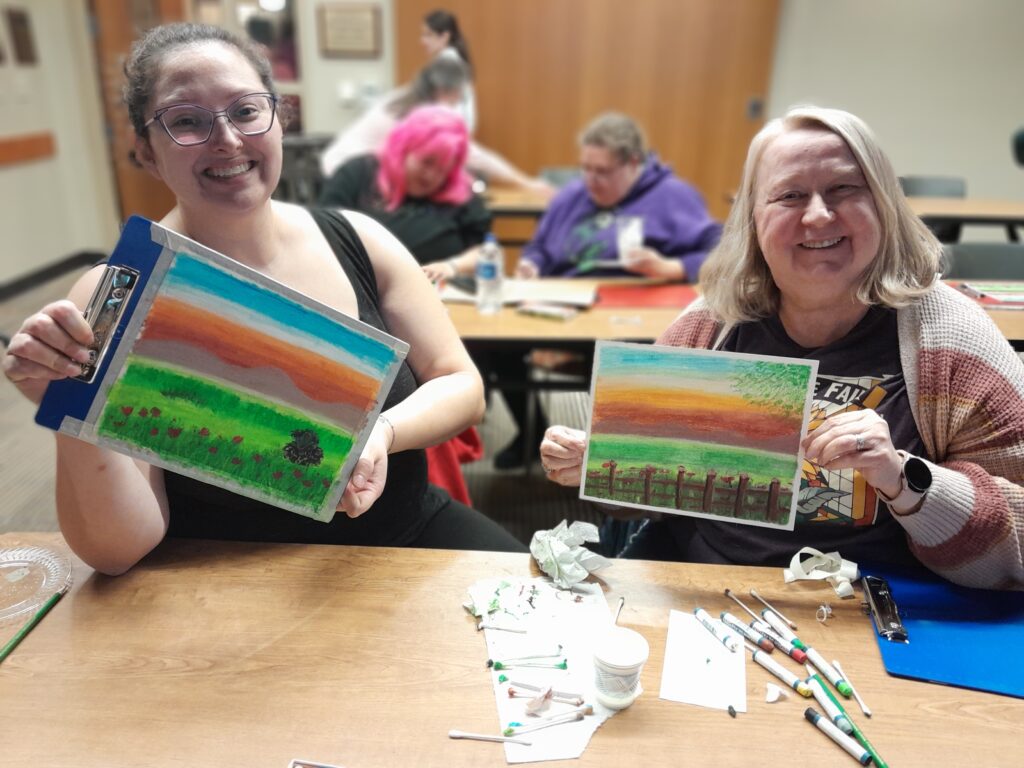 Two adults holding up their oil pastel art that they completed during an Art Nite Library program