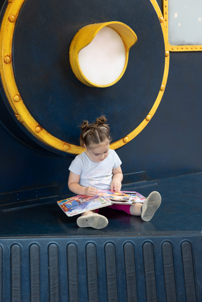 Little girl wearing a white shirt and red leggings reading a book on a bench shaped like a train